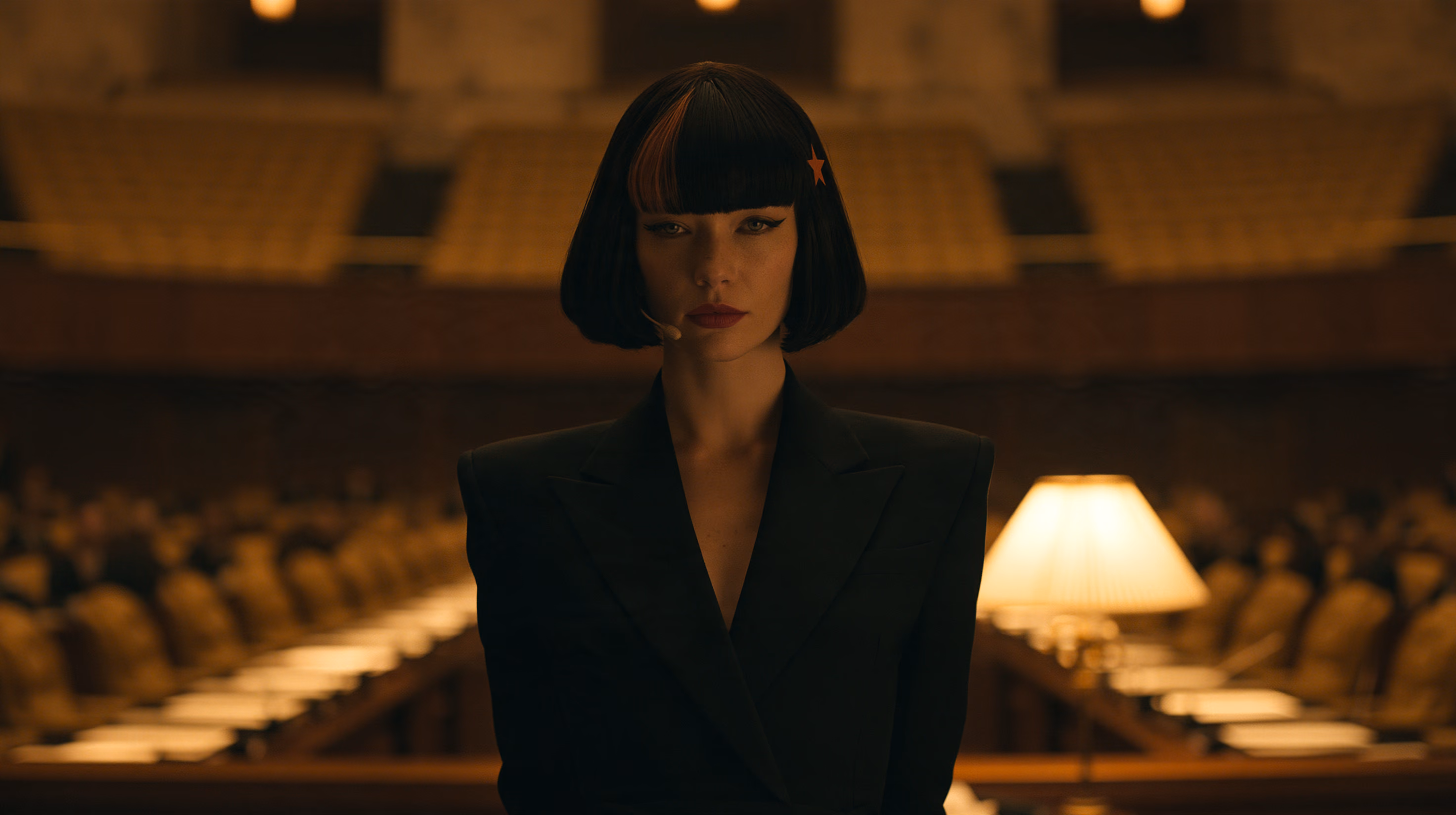 Claudia, the fan created character, standing in a dark council chamber in a black suit, a lamp glowing behind her
